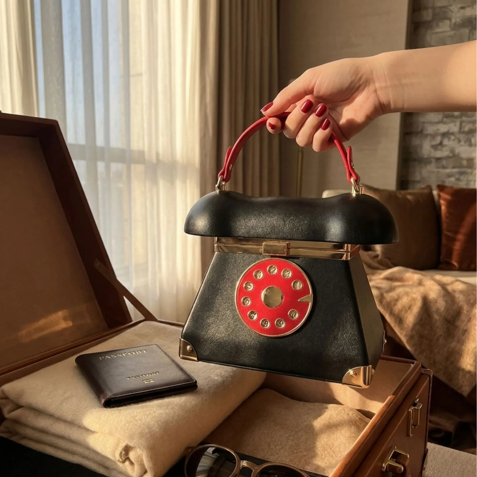 Dans un format 1x1, le sac est disposé à l'intérieur d'une valise de créateur ouverte, niché à côté d'une trousse de toilette en cuir assortie et d'un passeport élégant. Une main délicate, aux ongles vernis en rouge, tient le sac par sa poignée rouge, prête à le glisser dans un sac de voyage plus grand. La lumière dorée de l'après-midi pénètre à travers la fenêtre d'une chambre d'hôtel, évoquant le début d'un voyage stylé.